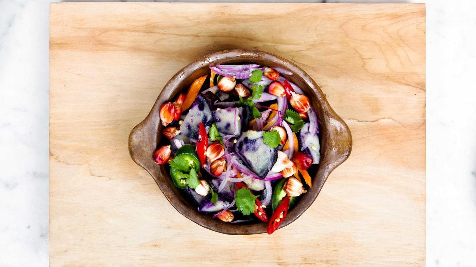 A close-up overhead shot of an earthenware bowl filled with a colorful salad featuring purple potatoes, red onion slices, vibrant red and green peppers, and toasted corn kernels, all garnished with cilantro leaves, resting on a light wooden cutting board.